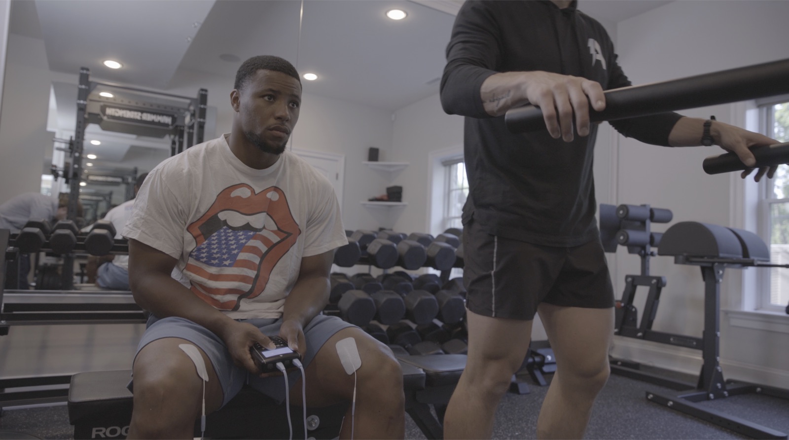 Saquon Barkley seated on a gym bench with Volta X electrode pads on both knees, holding the device controller during an Accelerate ACL training session